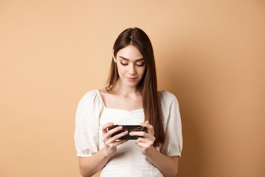 Pretty girl watching videos on smartphone, holding mobile phone horizontally and looking at screen, standing on beige background.