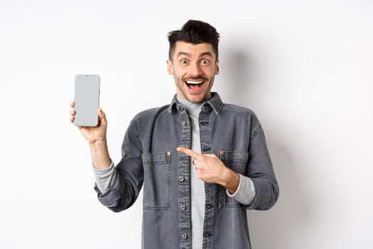 Excited handsome man showing empty smartphone screen and smiling, look here gesture, standing against white background.