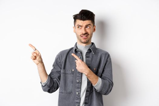 Skeptical and doubtful young man pointing fingers left at logo, look with dislike and confusion, standing on white background.
