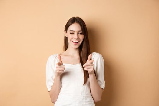 We need you. Smiling young woman pointing fingers at camera to beckon or invite people, standing on beige background.