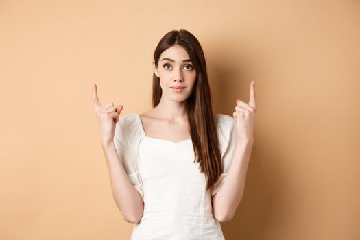 Hopeful romantic girl in white dress pointing fingers up, showing empty space for company logo, standing on beige background.