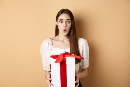 Image of surprised girl receive gift on Valentines day, looking with disbelief and holding present, standing on beige background.