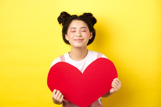 Cute teenage asian girl dreaming of true love, close eyes and showing big red heart cutout, waiting for soulmate on Valentines day, standing with romantic makeup on yellow background.