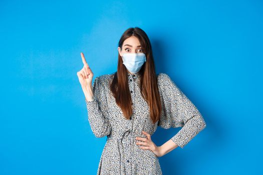 Healthy people and covid-19 pandemic concept. Excited woman in medical mask pointing at upper left corner, standing on blue background.