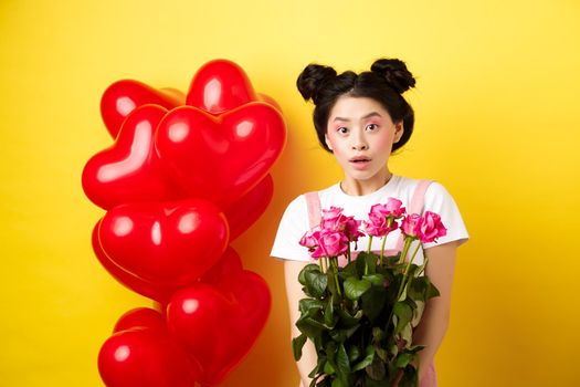 Happy Valentines day. Surprised asian girl receive bouquet of pink roses from lover, looking with awe at camera, standing near red heart balloons, yellow background.