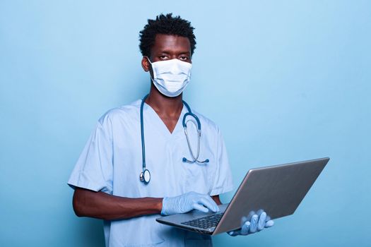 Healthcare specialist with laptop in hand looking at camera. Medical nurse with stethoscope and blue uniform wearing face mask against coronavirus pandemic. Assistant with gloves