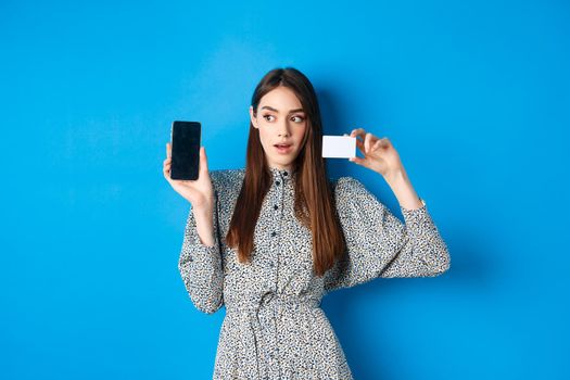 Online shopping. Pensive lady in dress showing empty phone screen and plastic credit card, order in internet, blue background.