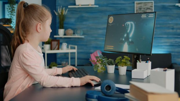 Young girl playing video games on computer after online school and homework. Gamer using shooting action play for entertainment and fun with keyboard and monitor. Child enjoying game