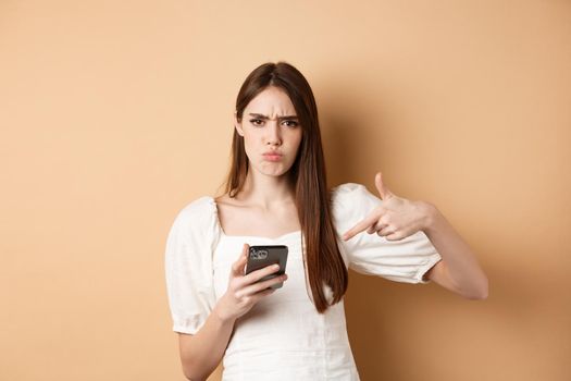 Online shopping concept. Angry frowning girl pointing at mobile phone screen, look disappointed, standing on beige background.