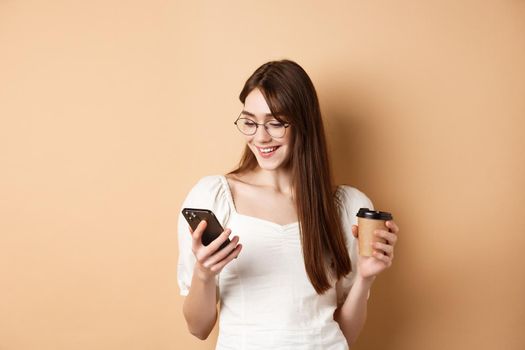 Happy girl chatting on phone and drinking coffee, wearing eyewear and white blouse, smiling at smartphone screen, beige background.