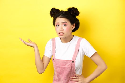 Confused asian teen girl with bright makeup, raising hand up clueless, looking puzzled, cant understand something, pointing at copy space, yellow background.