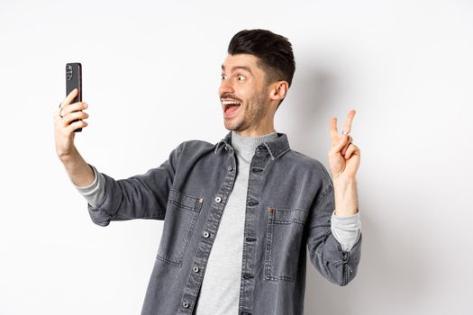 Profile of cheerful young guy taking selfie on smartphone, showing v-sign while make photo on mobile app, standing against white background.