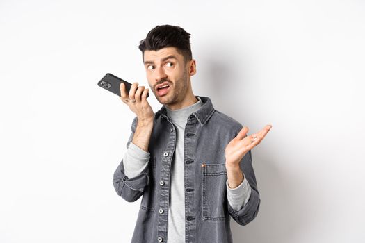 Stylish handsome man talking on speakerphone, record voice message with smartphone near lips, discuss something and looking aside at empty space, standing on white background.