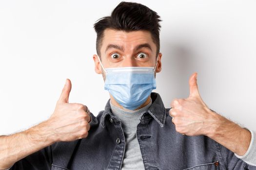 Covid-19, health and lifestyle concept. Close-up of excited positive guy in medical mask supports wearing medical mask during pandemic, showing thumbs up in approval.
