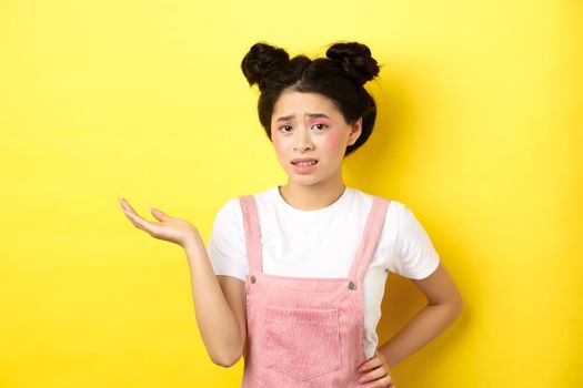 Confused teen asian girl with bright makeup and summer clothes, raising hand up and frowning puzzled, cant understand, yellow background.