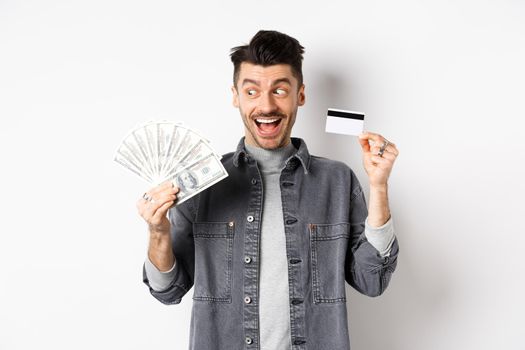 Excited guy holding plastic credit card and dollar bills, standing amused on white background.