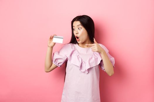 Excited asian woman pointing and showing plastic credit card, gasping amazed, concept of shopping.