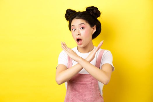 She says no. Asian beauty girl with makeup, showing cross sign, stop and reject offer, standing disappointed on yellow background.