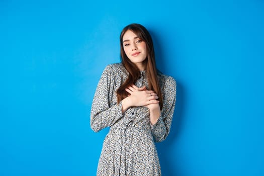 Romantic beautiful woman in dress holding hands on heart, smiling tenderly, keep sweet memories, standing on blue background.