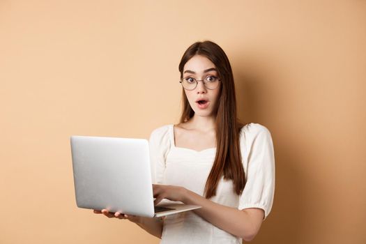 Surprised girl using laptop and gasping amazed, say wow, looking in glasses at camera, beige background.
