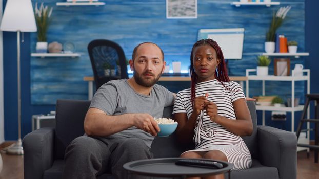 POV of married interracial couple watching scary movie together on couch. Multi ethnic husband and wife eating popcorn being afraid while looking at camera on living room sofa.