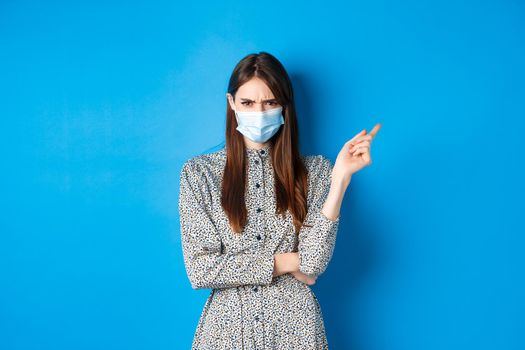 Covid-19, social distancing and healthcare concept. Angry young woman in medical mask frowning, pointing right at bad thing, condemn non-maskers, blue background.