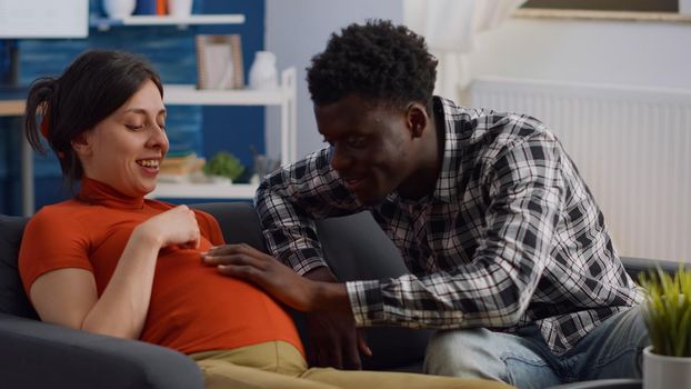 Pregnant interracial couple sitting on living room couch. African american husband touching baby bump and talking to child while caucasian mother relaxing on sofa and laughing