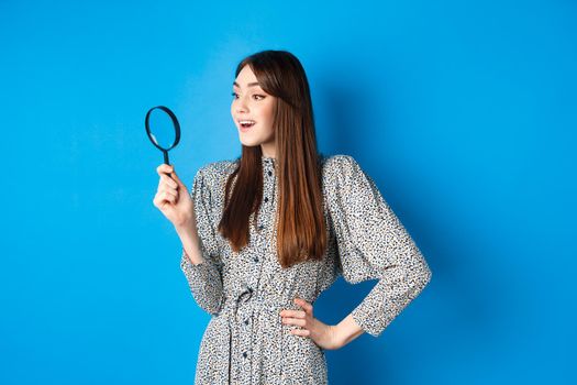 Excited girl looking left with magnifying glass, found interesting promo, investigating or searching, standing on blue background.