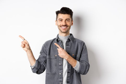 Handsome smiling man pointing fingers left, showing logo and looking cheerful at camera, advertising on white background.