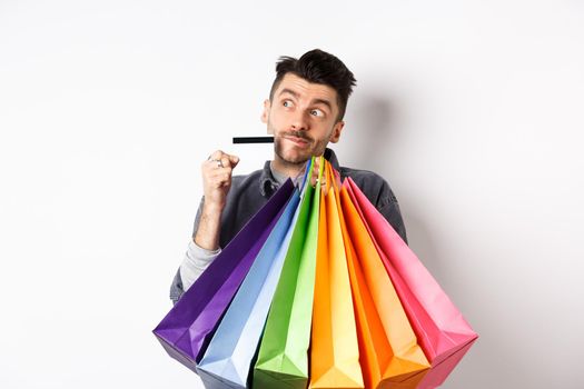 Dreamy guy thinking of shopping, holding colorful bags with purchased items and standing thoughtful with credit card, white background.