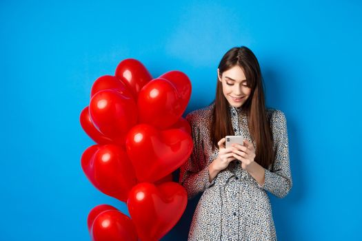 Valentines day. Smiling woman in dress standing near red hearts balloons and looking at smartphone, standing on blue background.