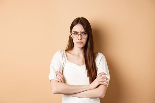 Angry and offended girl in glasses frowning, cross arms on chest and sulking, standing defensive on beige background.