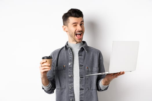 Happy male worker drinking coffee and work with laptop, winking at camera, standing on white background.