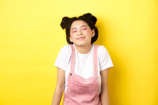 Romantic asian girl with cute pink makeup and summer clothes, close eyes and smiling dreamy, standing on yellow background.