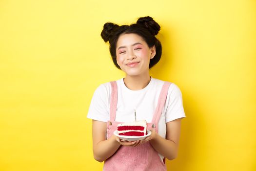 Happy asian birthday girl with bright makeup, blowing candle on cake, making wish, standing on yellow background.