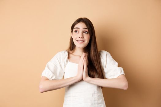 Hopeful young girl begging god and looking dreamy at upper left corner, praying or making wish, standing on beige background.