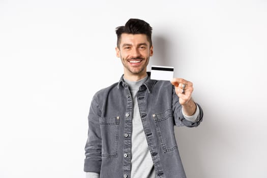 Handsome stylish guy showing plastic credit card and smiling, satisfied client standing on white background.