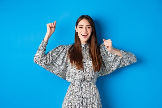 Cheerful young woman in dress raising hands up and smiling excited, triumphing over winning prize, celebrating victory, standing on blue background.