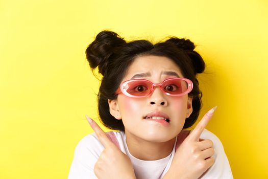Summer fashion concept. Headshot of worried indecisive asian girl, pointing fingers sideways and showing two ways, cant choose, wearing sunglasses.