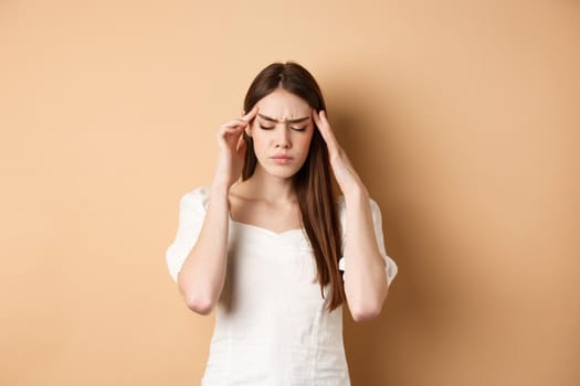 Young woman having headache, touching head temples with closed eyes and tensed face, standing with painful migraine on beige background.