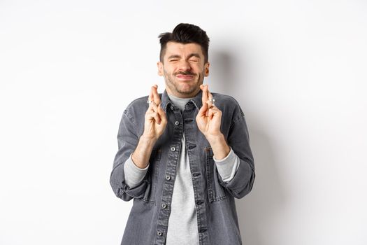 Hopeful young man standing nervous with closed eyes and fingers crossed, making wish, begging god for dream come true, waiting news, standing on white background.