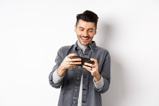 Handsome young man playing mobile video game and smiling at screen, standing against white background.