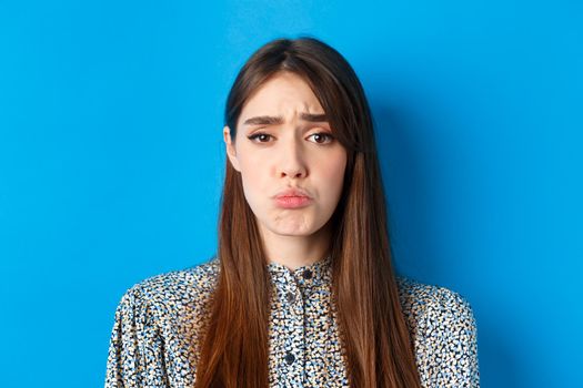 People emotions concept. Sad cute girl begging for something, pucker lips and frowning, whining asking please, standing on blue background.