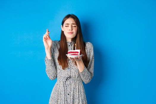Hopeful birthday girl wish for dream come true, cross fingers with eyes closed, holding cake with candle, blue background.