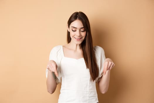 Smiling beautiful female model looking and pointing down with pleased happy face, showing something romantic, standing on beige background.