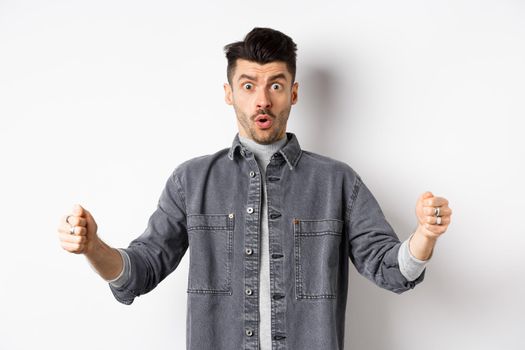 Confused man holding big size object and looking shocked, shaping large thing, standing on white background.