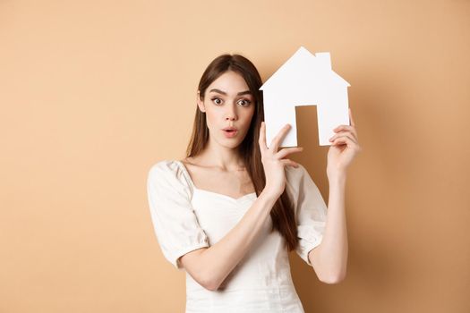 Real estate. Excited young woman showing house cutout and looking at camera, renting property, standing on beige background.