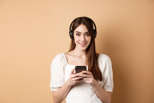 Pretty girl in headphones smiling at camera, listening music and using mobile phone app, beige background.