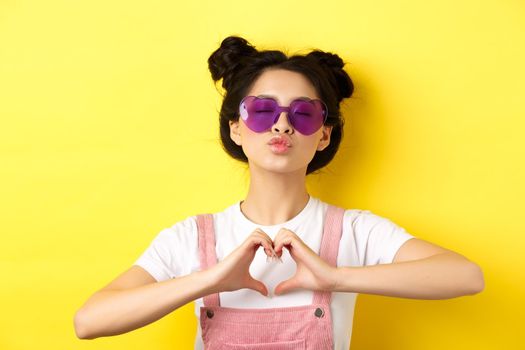 Summer and fashion concept. Cute glam girl with sunglasses and hairbuns, showing heart sign and pucker lips for kiss, I love you gesture, yellow background.