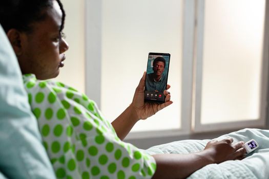 Young patient of african ethnicity talking on video call conference with smartphone while sitting in hospital ward bed. Black girl using online internet for telecommunication with man